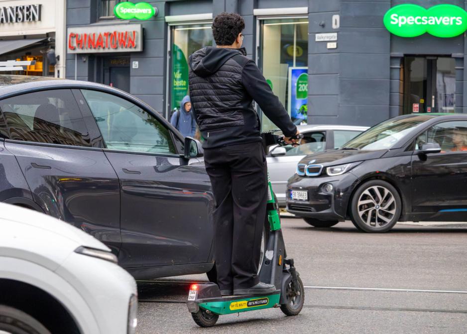 En ung mann eller tenåring kjører en elsparkesykkel på veien, mellom flere biler. Bilde er tatt slik at man ser han bakfra.