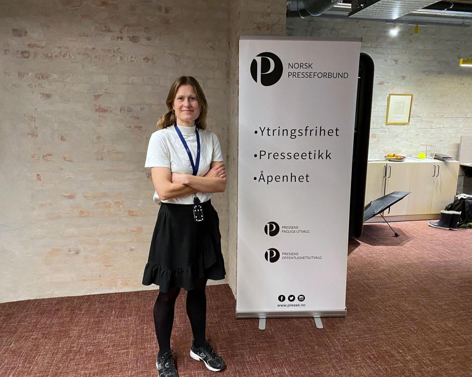 Kristine Foss står inne på kontoret sitt ved siden av en plakat fra Norsk Presseforbund som sier "Ytringsfrihet, Presseetikk, og Åpenhet"