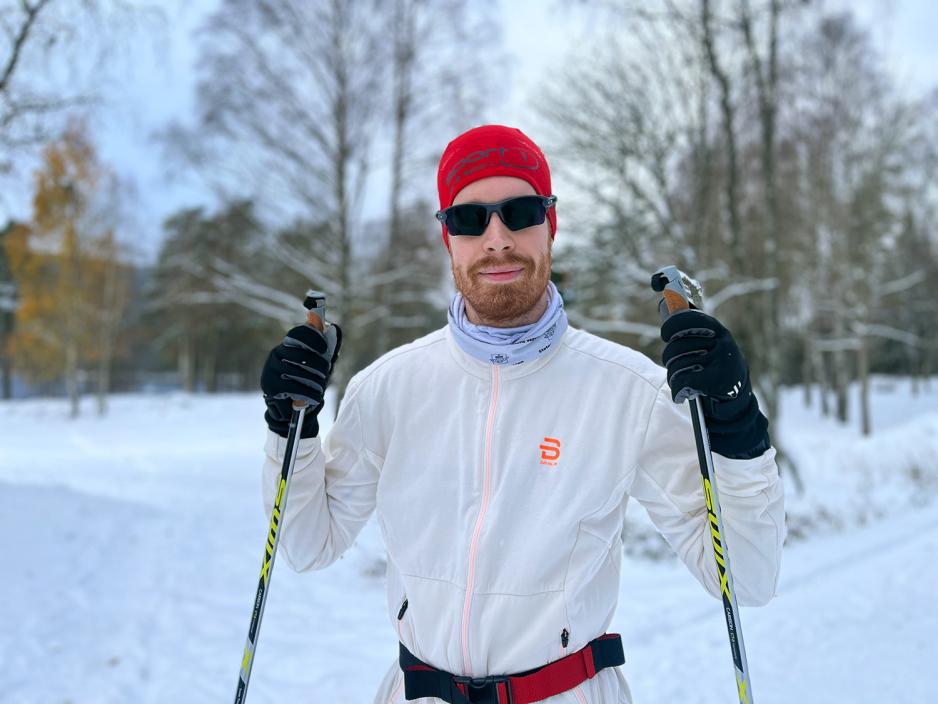 Mann med rød lue, solbriller, en tynn treningsjakke og en stav i hver hånd.