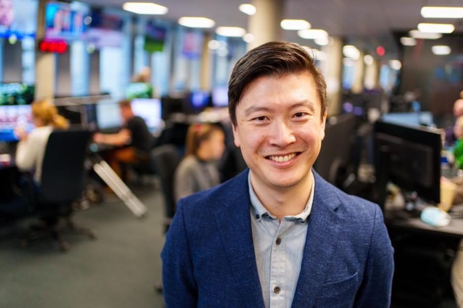 Portrett av Derek Bjølgerud. Han står i dressjakke og skjorte, og smiler til kamera. Han står på nyhetsgulvet i TV2s lokaler.