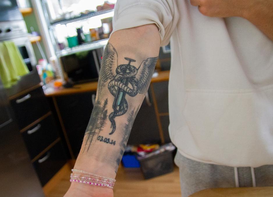 Tatovering på underarm, av en sprøyte med en slange rundt. På toppen er det vinger. Under sprøyten er det en dato. 