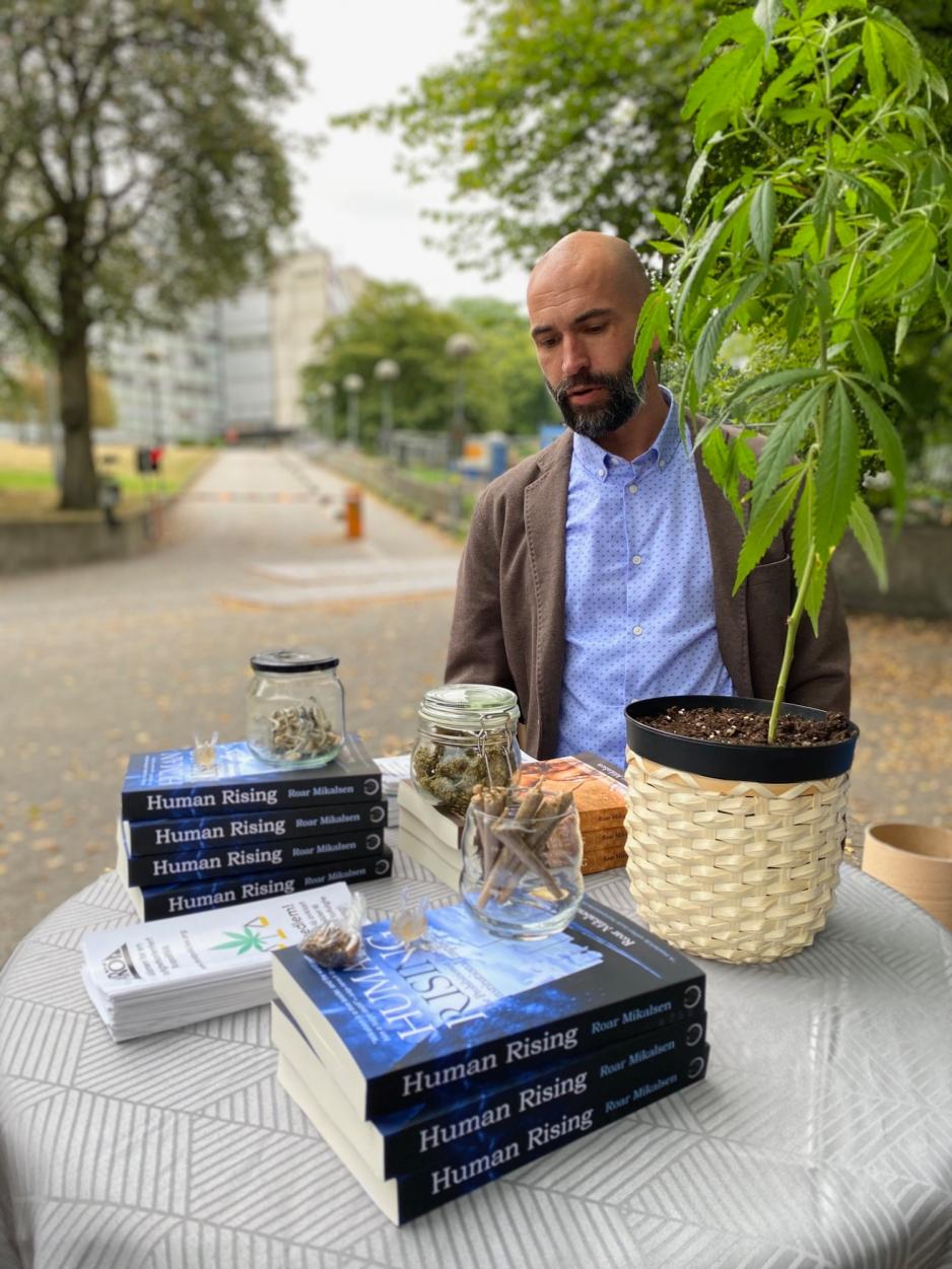 Roar Mikalsen står ved bord med krukker med stoff, bøker og en cannabisplante. 