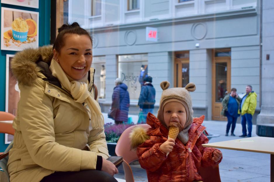 Liten gutt med bamselue og orange jakke spiser is, mens moren, en dame med mørkt hår og lyst skjerf, sitter på huk ved siden av