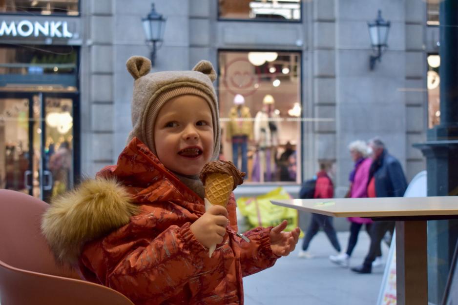 Liten gutt med bamselue og orange vinterakke spiser is på en isbar i Karl Johans gate. Utenfor er gaten travel. 