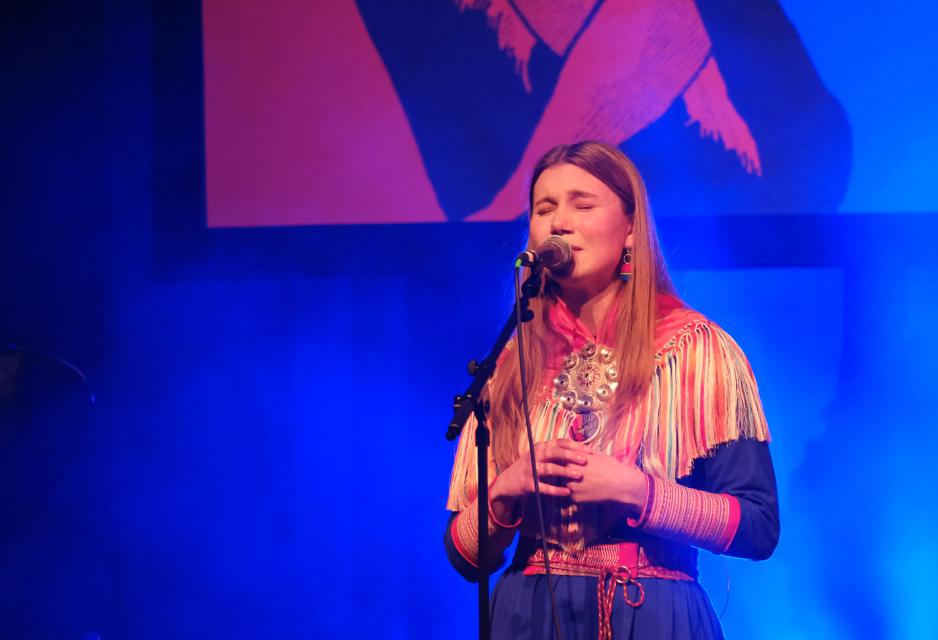 Ella Marie Hætta Isaksen synger på støttekonserten. Hun er i kofte, og det er blått lys bak henne. 