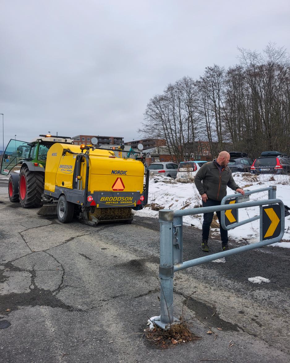 I bakgrunnen ser man en grønn traktor med gult feieslep, og i forgrunnen åpner sjåføren en bom som han skal kjøre gjennom.