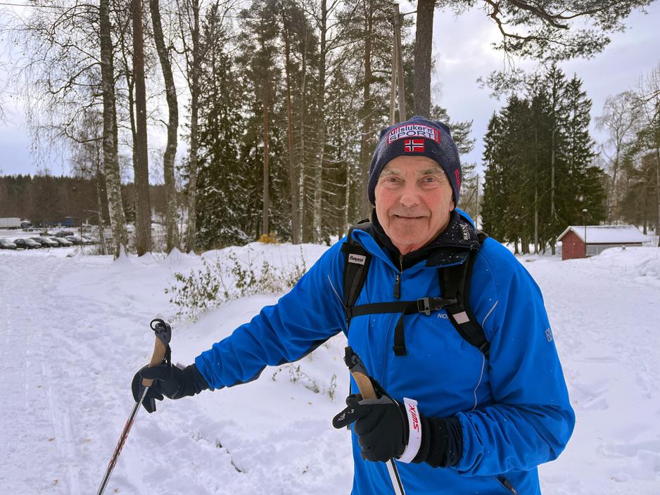 Eldre mann med med en mørk lue, en blå jakke, sekk på ryggen og har på skiutstyr