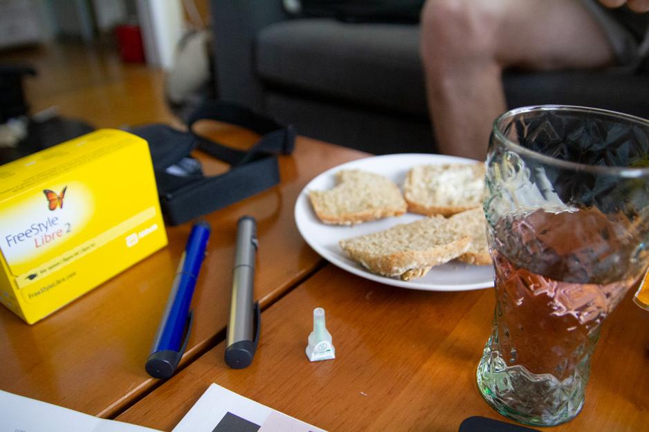 På et brunt bord ligger det to insulinpenner. Ved siden av står et glass saft og rundstykker på en tallerken. 