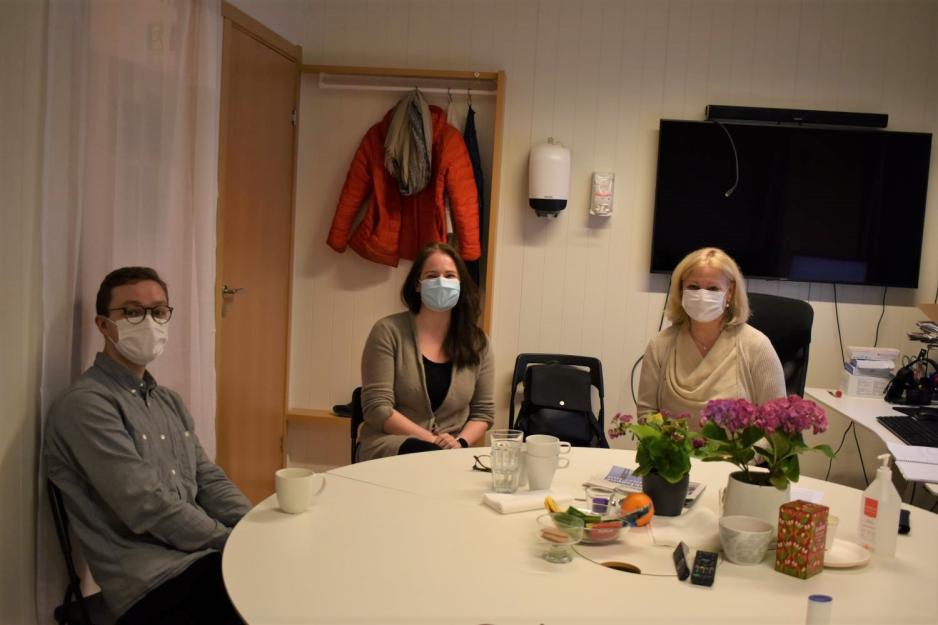 Rundt et rundt bord på et kontor sitter en ung mann, en ung mørkhåret kvinne og en kvinne i 40-50 årene. Det er en typisk møtesituasjon, der de har frukt og pepperkaker på bordet. I bakgrunnen står en datamaskin. 