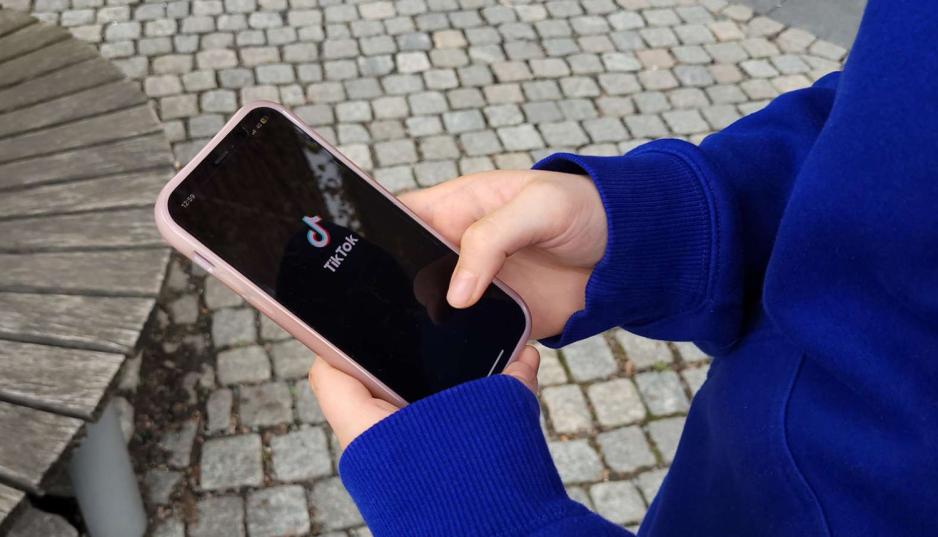 Bildet viser en jente som ser på telefonen sin. Hun har TikTok oppe og logoen til TikTok vises på skjermen. Jenta har på seg blå genser og står ute.