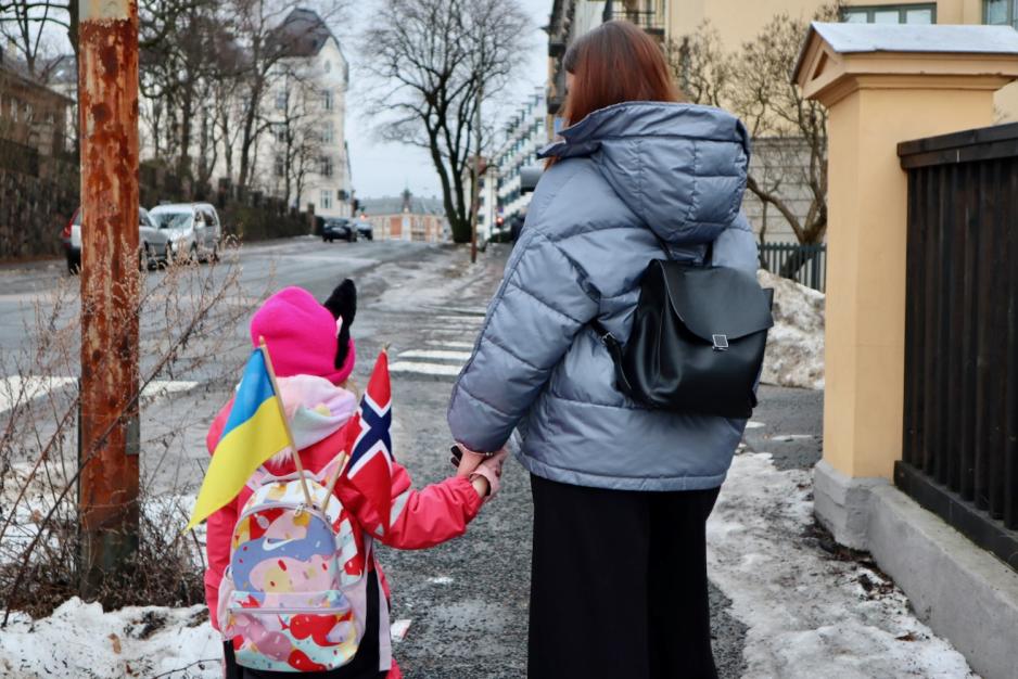 Mor og datter bakfra som holder hender. Et Ukrainsk og et norsk flagg stikker ut av sekken til datteren.