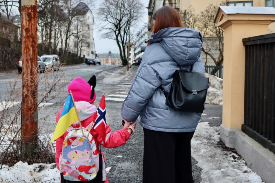 Mor og datter bakfra som holder hender. Et Ukrainsk og et norsk flagg stikker ut av sekken til datteren. 