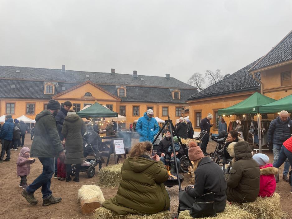 Bildet viser en gjeng som sitter og spiser mat på høyballer rundt en bålpanne. I bakgrunnen ser man den oransje hovedbygningen på Bogstad Gård. 