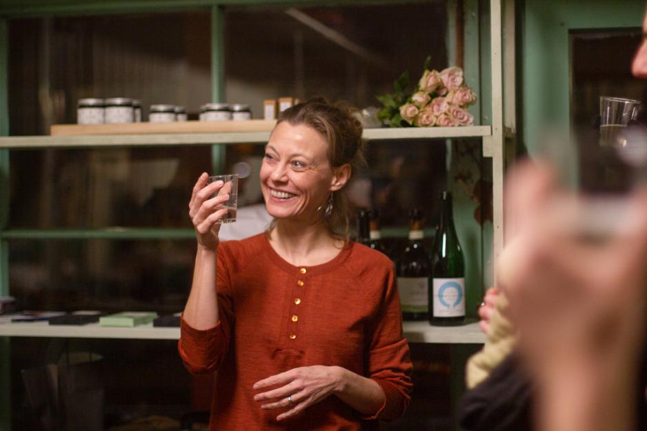 Frøydis skåler til menneskene som kom på åpningen til det nye lokalet. Hun er ikledd en rød topp fra Fogg og smiler. Man ser glass i forgrunnen av bildet, og hyller i bakgrunnen.