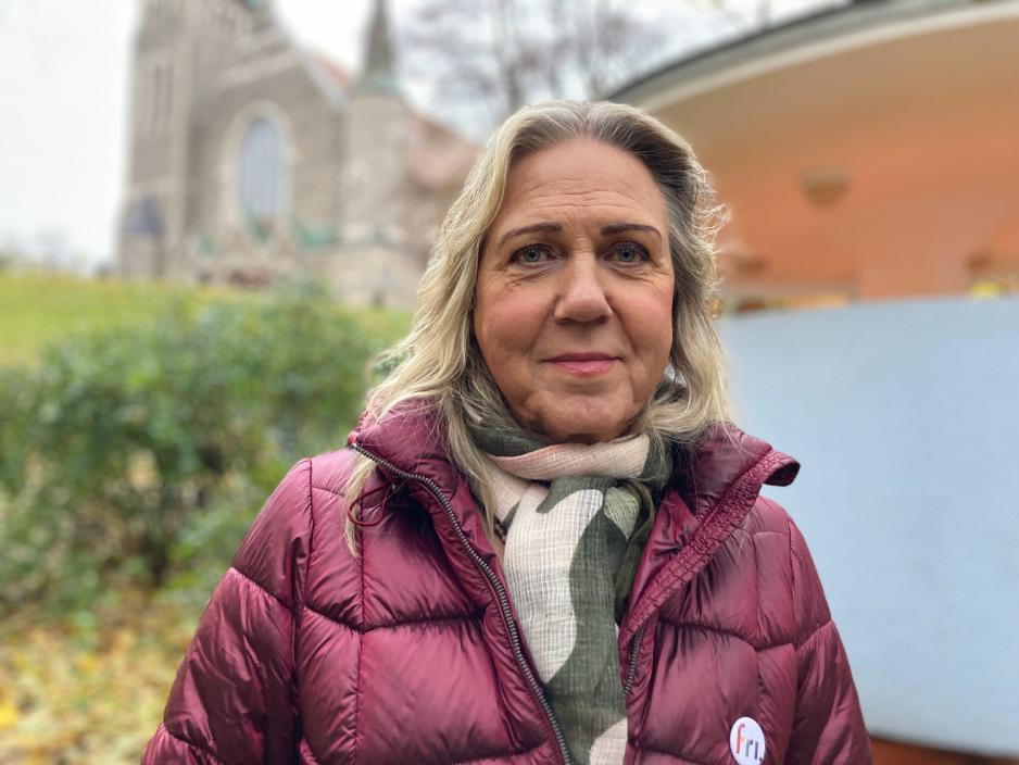 Jan Elisabeth Lindvik smiler foran Kjærlighetskarusellen og en kirke. Rundt Lindvik er det trær og løv i høstfarger.