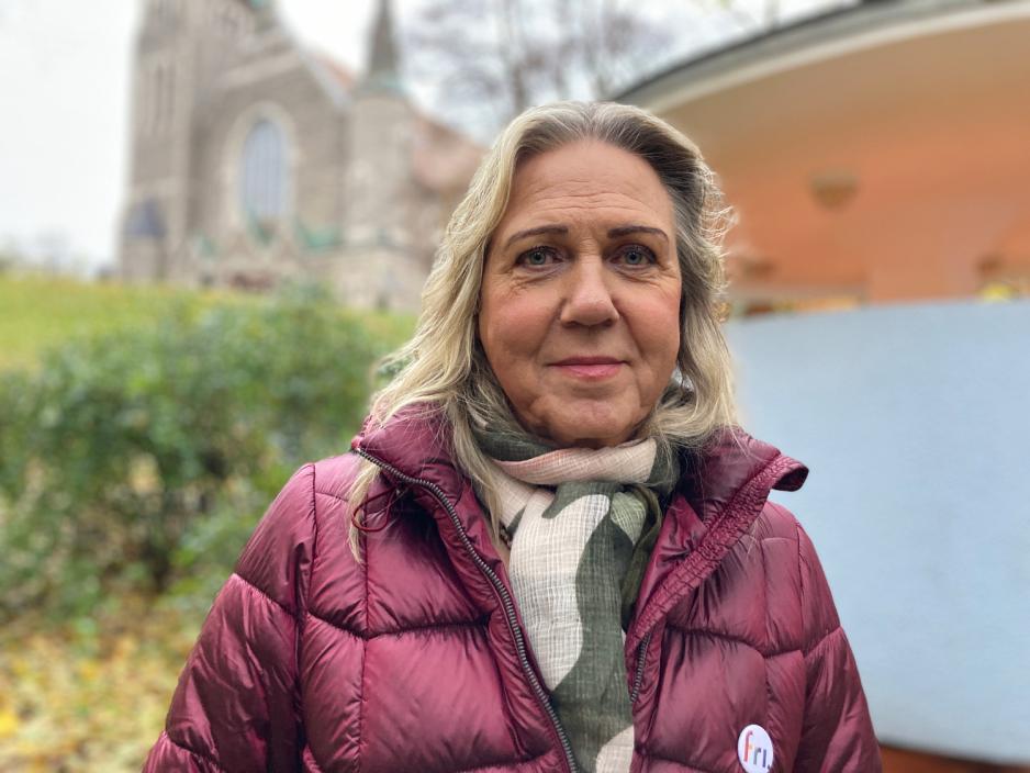 Jan Elisabeth Lindvik smiler foran Kjærlighetskarusellen og en kirke. Rundt Lindvik er det trær og løv i høstfarger. 