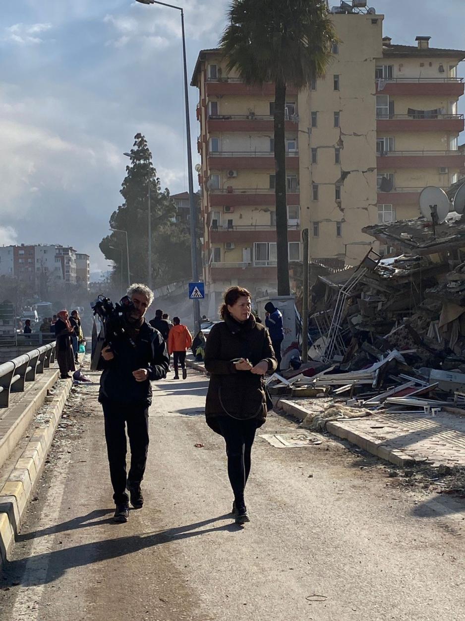 En fotograf med videokamera og en journalist går på sandete vei ved siden av et rast bygg