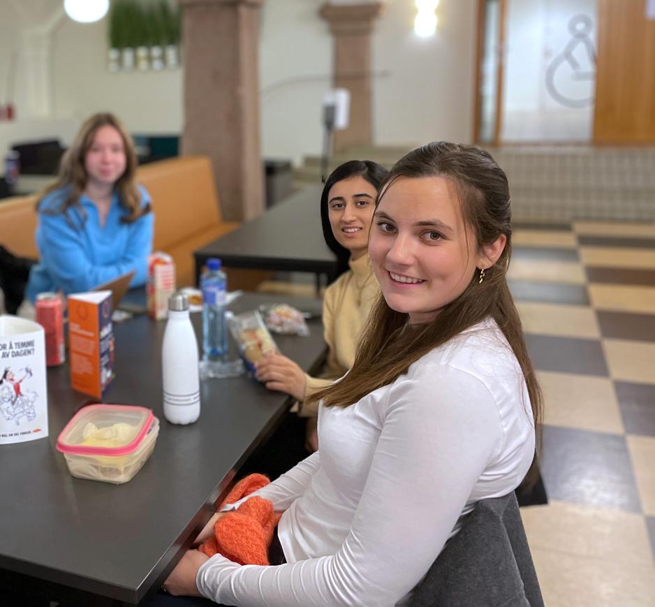 Studentene Karoline og Simran smiler til kameraet. De spiser egen mat i kantina.