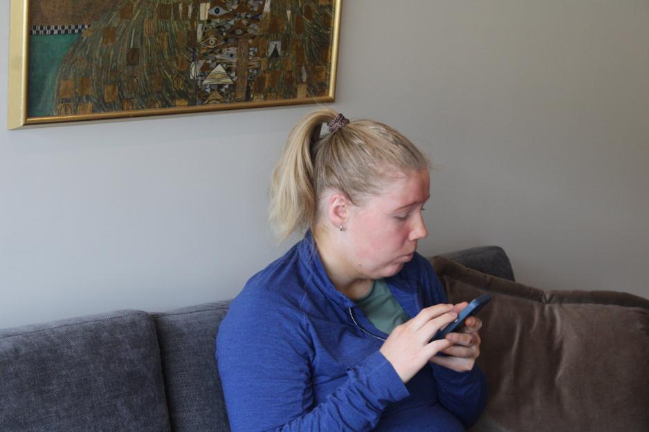 Kristin Hauglid sitter i sofaen, med telefonen nærme ansiktet. Hun justerer hastigheten på på skjermleseren på telefonen.