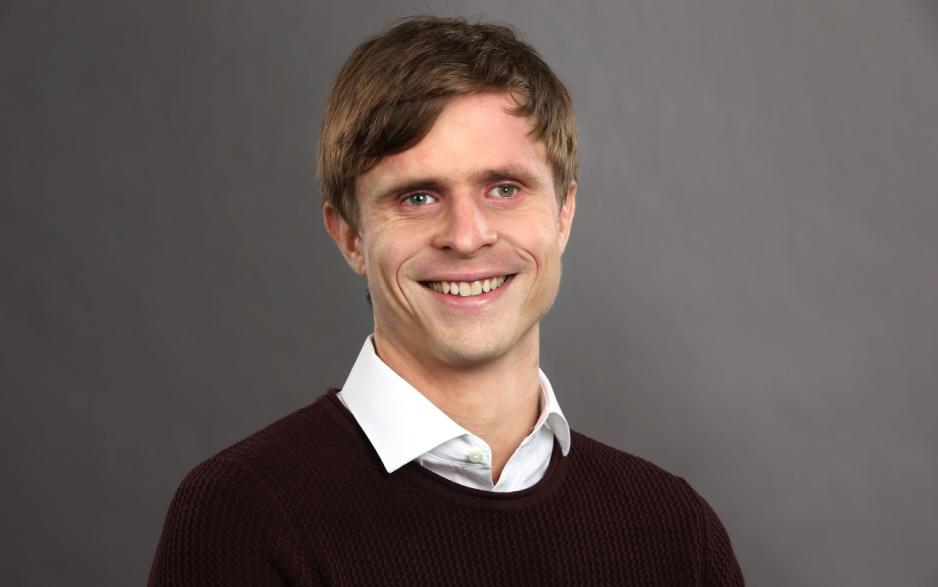 Portrettbilde av Kristoffer Lium fra NRK. Han står foran en mørk grå bakgrunn, og har på seg hvit skjorte og mørkerød genser. Han smiler inn i kamera.