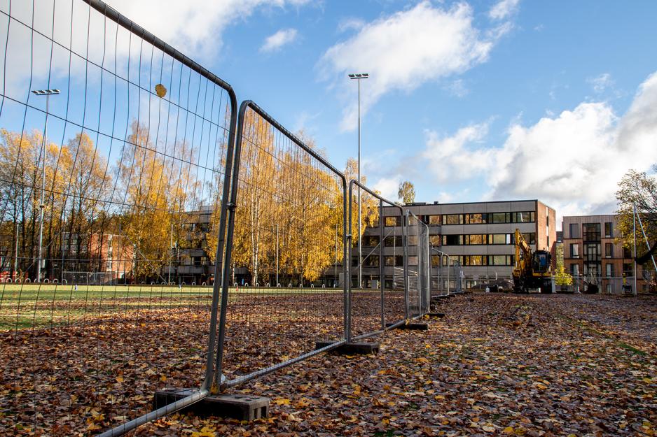 Kunstgressbane med mye løv og et byggegjerdet med en gravemaskin og et boligbygg i bakgrunnen