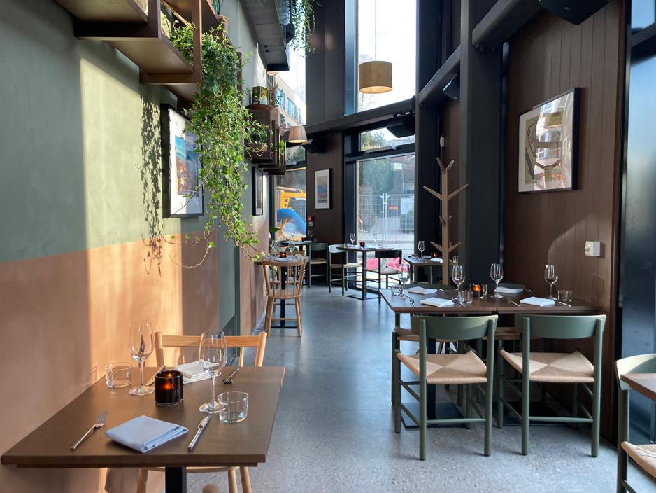 Bildet viser en del av restaurantens lokale. Det er planter, olivengrønne og rustenoransje vegger i tillegg til mye lys og sitteplasser i tre. På bordene er det lys. 