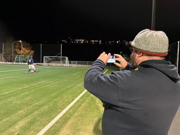 Marius Helgå bruker mobilen til å filme en hendelse på fotballbanen i bakgrunnen.