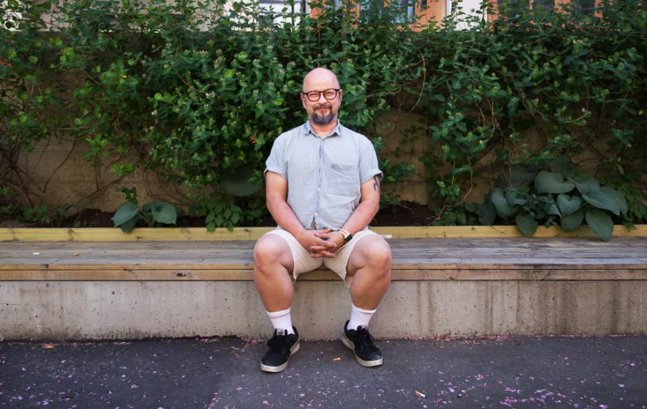 Portrett av Mikael Scott Bjerkeli hvor han sitter på en benk foran en busk. Hendene er foldet i fanget. 