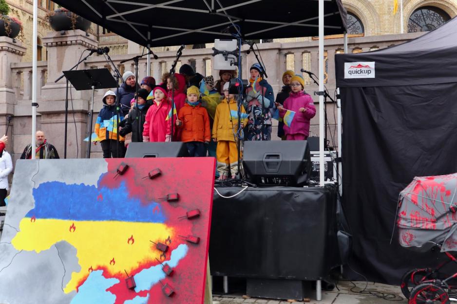 mange barn på en scene som holder det ukrainske flagget. de står foran stortinget. foran dem er et maleri av Ukraina og Russland, der russland er rødt og invaderer Ukraina.