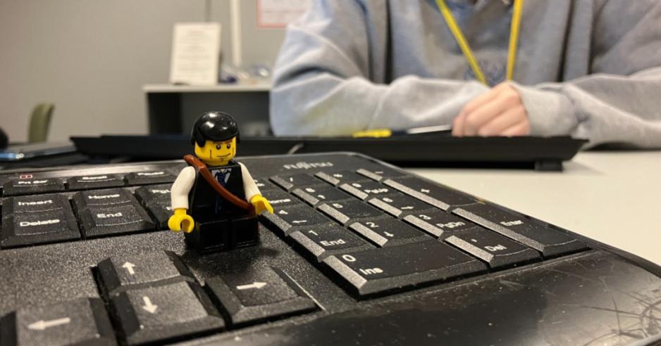 Legomann sittende på et tastatur (oppå et bord), med overkroppen til en dame og et annet tastatur synlig i bakgrunnen.