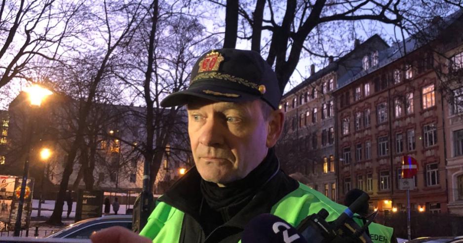 Politiets innsatsleder omringet av journalister, de er ikke synlige, bare mikrofonene/telefonene deres. I bakgrunnen ser man bygget det brenner i, dog ikke vinduet til brannleiligheten.