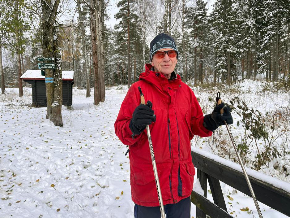 Eldre mann med rød anorakk, mørk lue, solbriller og en stav i hver hånd. 