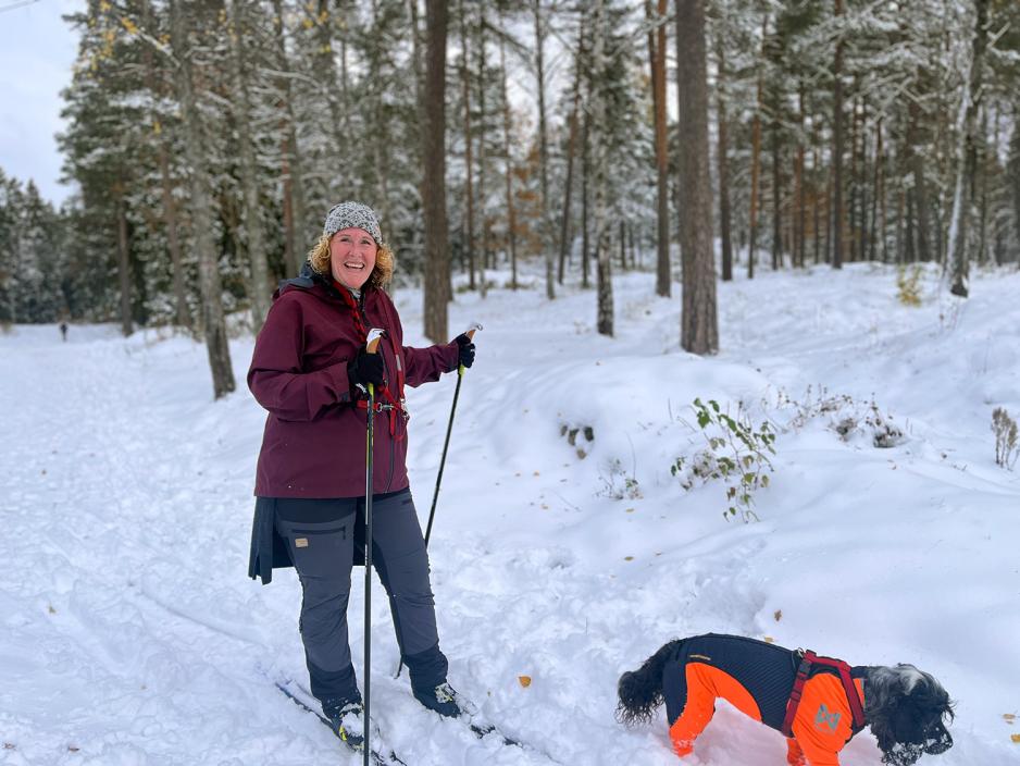 Voksen kvinne med burgunerrød jakke, grå lue og svart bukse. Hun er på skitur med hunden sin, en liten, sort og grå hund. 