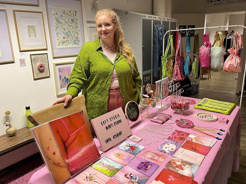 Fotograf og designer Sara Tonning bak sin markedsbod dekket med rosa duk, med postkort, smykker og et stativ med egenproduserte håndvesker i sterke iøyenfallende farger.