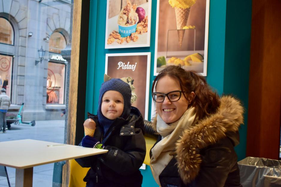 Glad dame med svarte briller og mørkt hår sitter ved siden av sønnen sin på omtrent to år. Han har mørke klær og spiser på en kule sjokoladeis i kjeks. 