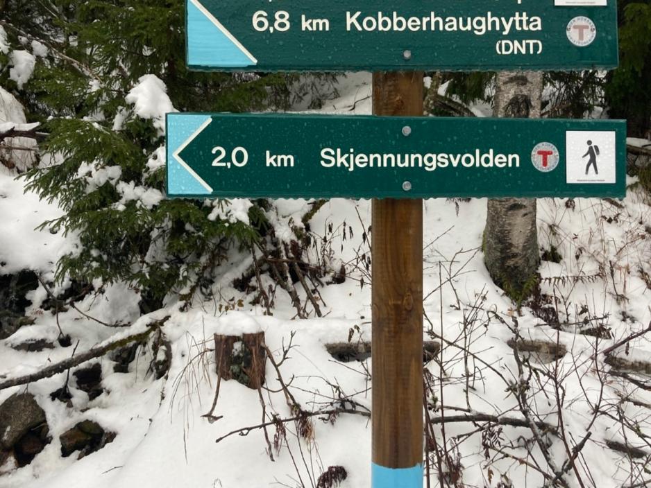 Et skilt som blant annet peker mot Skjennungsvolden og som sier at det er to kilometer dit. Det er snø i bakgrunnen. 