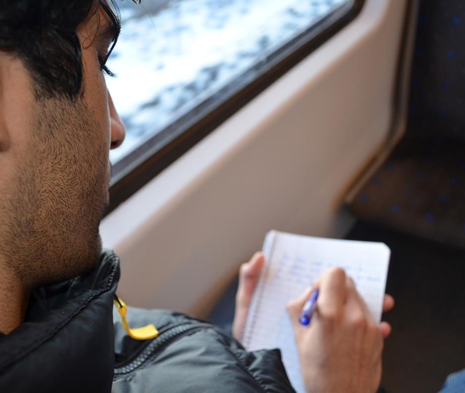 Person har et hodet med fokus på og sitter og skriver på t-banen. Blokken har skriver på er ikke i fokus. Vi ser over skuldrene hans