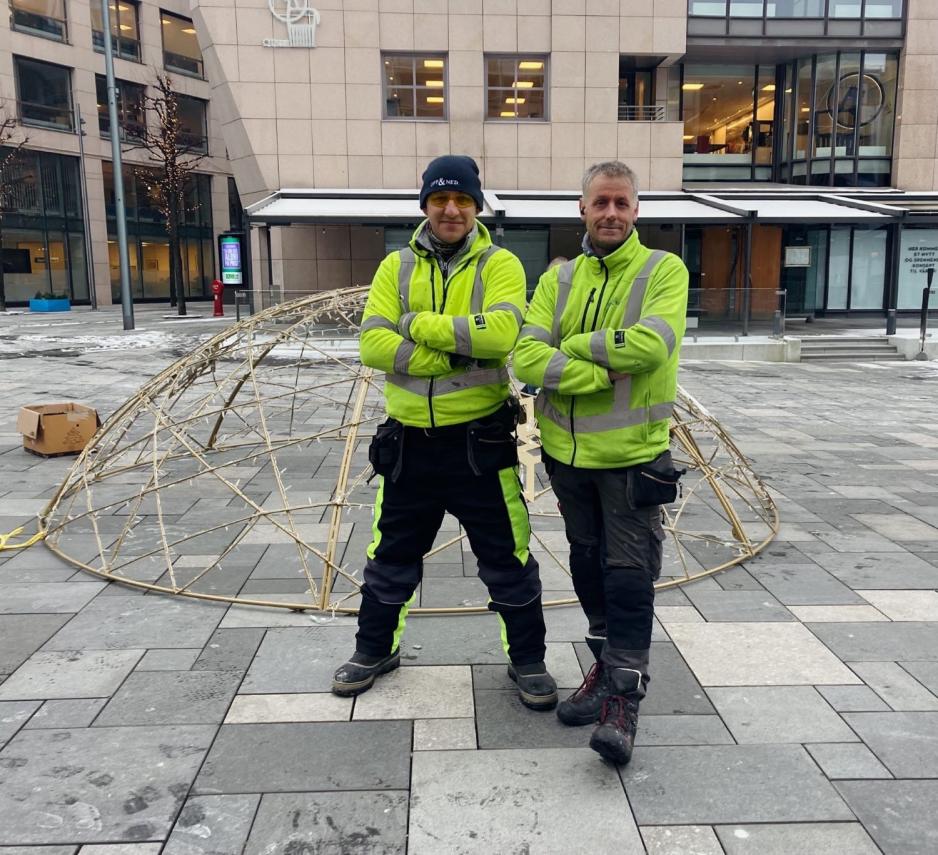 To menn i grønne jakker står foran den demonterte julekulen på Bryggetorget.