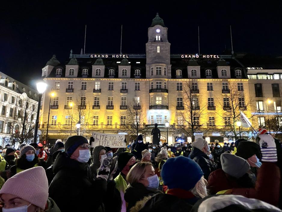 En godt påkledd folkemengde står tett i tett med munnbind på. Noen av demonstrantene hever et banner det står "lite lys og lite varme" på. 