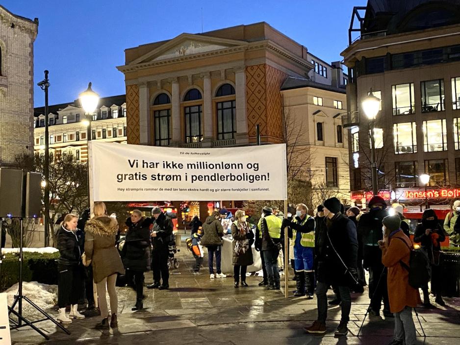 To menn holder et stort banner med teksten "Vi har ikke millionlønn og gratis strøm i pendlerboligen". Flere demonstranter vrimler rundt dem. 