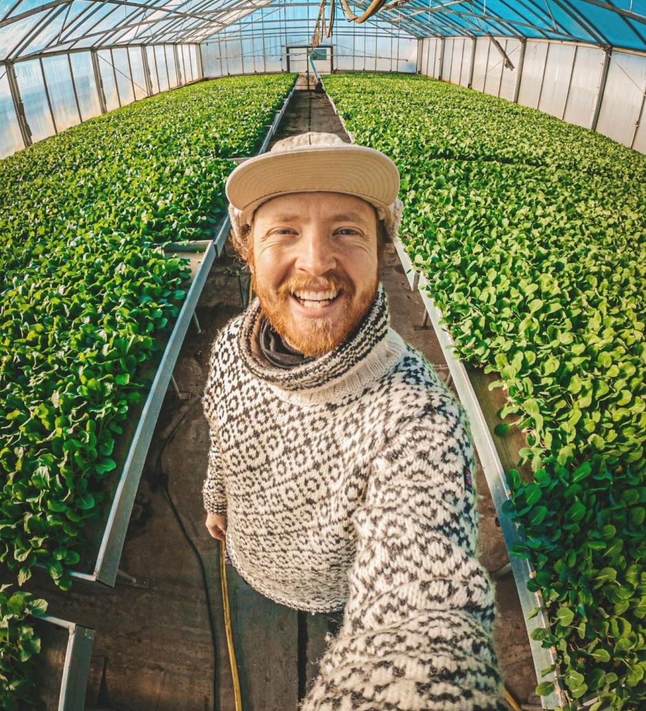 En mann smiler mot kamera, bak seg har han masse grønne planter. 