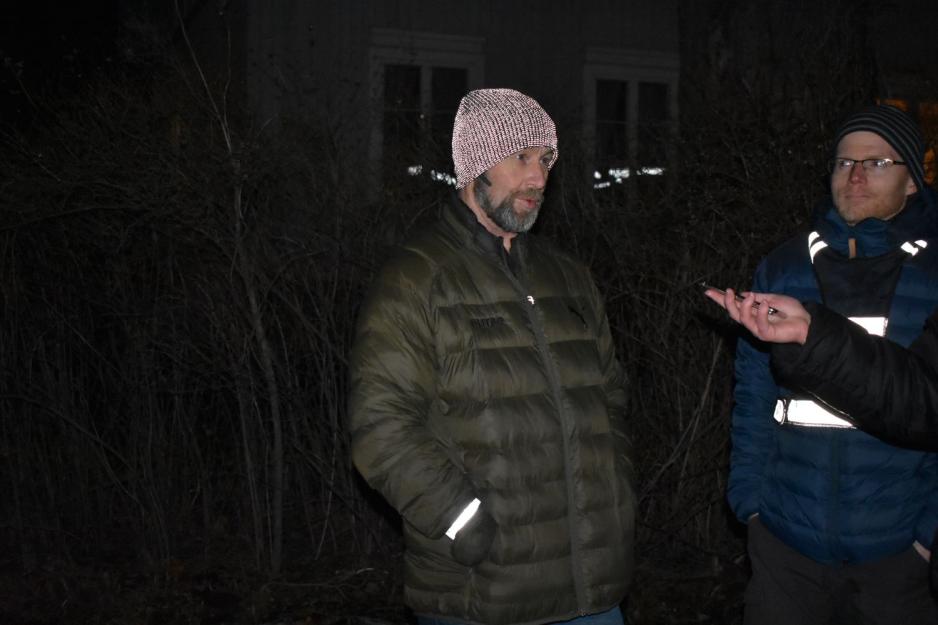 En mann med reflekslysende lue står med boblejakke og hendene i lomma. Til høyre i bildet står en mann med rødlige skjeggstubber og lytter interessert til det han sier. Det er mørkt.