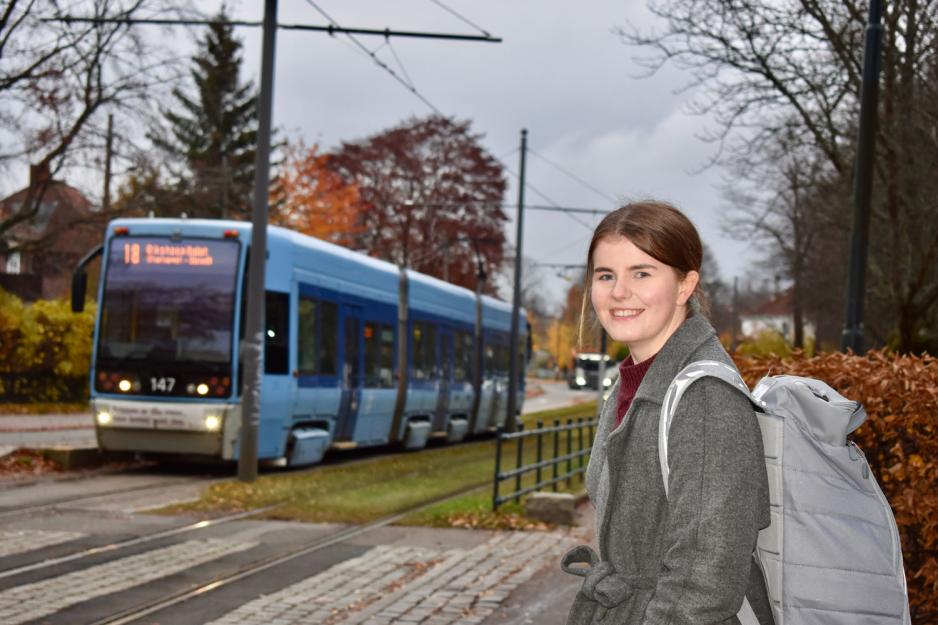 Ung kvinne med mørkt hår og grå kåpe venter på trikken. En trikk kjører i bakgrunnen. 