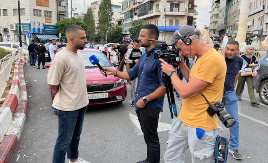 Journalist avbildet mens han intervjuer en annen mann, mens de begge blir filmet av en annen mann.