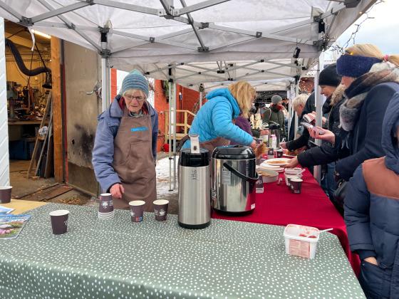 To kvinner fra venneforeningen botanisk hages venner som står bak et bord. den bakerste gjør klar en vaffel. kvinnen nærmest kamera er på vei å fylle på kaffe. utenfor bode er det folkerikt.