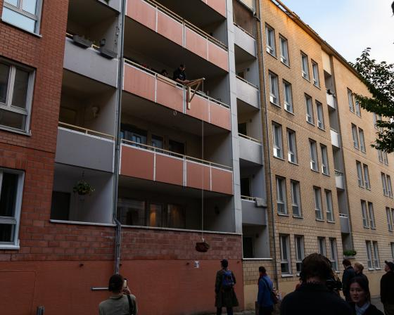 En blokk med en balkong med en hjemmelaget kran, som heiser ned en kurv med pizza esker. Det står en menneske samling og venter på gata.