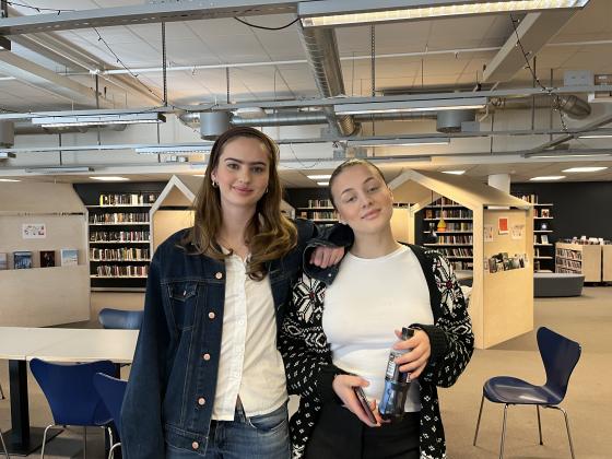To unge jenter står ved siden av hverandre på biblioteket og smiler