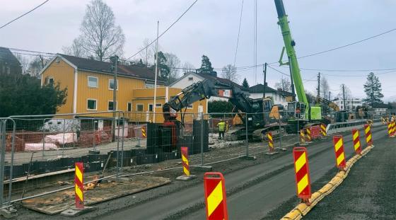 En høy grønn maskin til høyre. Foran denne maskinen står det en gul og sort gravemaskin. Rundt maskinene er det et langt gjerde av metall. I bakgrunnen ser man et gult hus. Foran maskinene er det en smal grusvei.