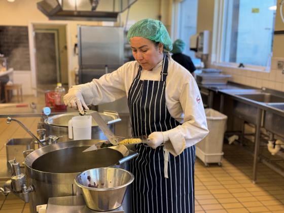Andrea står på et kjøkkenet ovenfor en kjele og skyver grønnsaker fra en fjøl over i kjelen. Hun har på forkle og hårnett og konsentrerer seg på matlagingen. 