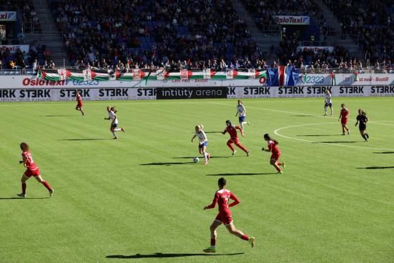 To fotballlag spiller mot hverandre på en kunstgressbane. Det palestinske laget har rød drakter. Vålerenga sitt lag har hvite drakter. Det er tilskuere på tribunen.  